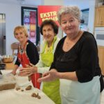 Tri starejše ženske v predpasnikih stojijo ob mizi in oblikujejo piškote v obliki polmeseca, razporejene na pekaču.