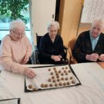 Tri starejše osebe sedijo ob mizi in ročno oblikujejo majhne kroglice testa, razporejene po pekaču.