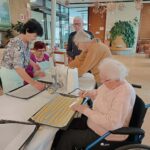 Skupina starejših oseb skupaj pripravlja piškote; ena oseba uporablja strojček za testo, druge pomagajo pri razporejanju trakov testa na pekač.