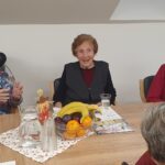 Starejše gospe sedijo za mizo, pred njimi so sadje, kozarci in skodelice; ena izmed njih z živahno kretnjo pripoveduje, druge pozorno poslušajo.