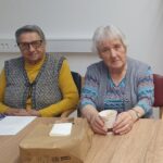 Dve starejši gospe sedita druga ob drugi, ena drži skodelico kave; obe gledata proti kameri z umirjenim izrazom.