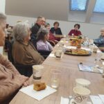 Večja skupina starejših oseb sedi okoli dolge mize, pred njimi so pijača in sladice; sproščeno se pogovarjajo in družijo.