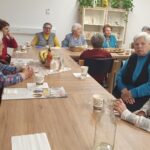 Starejši udeleženci sedijo okoli mize, na kateri so pijača, pecivo in sadje; med pogovorom se smehljajo in izmenjujejo misli.