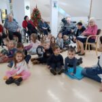 Otroci sedijo na blazinah na tleh, za njimi stanovalci doma starejših na stolih in vozičkih, v ozadju okrašena novoletna smreka.