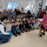 Starejša gospa bere pravljico otrokom, ki sedijo na tleh, okoli njih pa sedijo in poslušajo stanovalci doma starejših.