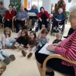 Otroci sedijo na tleh in poslušajo pravljico, za njimi stanovalci doma starejših, nekateri na invalidskih vozičkih.