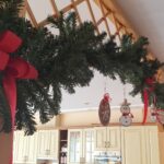 Božični okras iz zelenja z rdečimi trakovi in visečimi okraski nad prehodom v kuhinji.