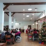 Več stanovalcev doma za starejše sedi za mizami v jedilnici in sodeluje v praznični tomboli; prostor je okrašen z božično jelko.