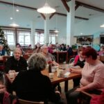 Skupina stanovalcev, zbranih okoli mize, rešuje listke za tombolo; na mizi so papirnati kozarci in pisala.