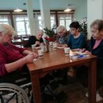 Pet starejših stanovalk sedi za mizo in izpolnjuje tombolske listke v svetlem skupnem prostoru doma.