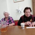 Dve stanovalke sedita za mizo z vrčem pijače in tombolskimi listki ter se nasmihata v praznično okrajenem prostoru.