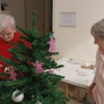 Dve stanovalki okrašujeta manjšo novoletno smrečico z okraski v skupnem prostoru doma.