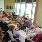 Skupina stanovalcev sedi ob dolgi mizi in ustvarja praznične venčke; pred njimi so material, okraski in zelenje, v ozadju pa svetel prostor z velikimi okni.