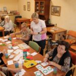 Skupina stanovalcev pri mizi ustvarja praznične motive na papir; pred njimi so barve, čopiči in kartončki z narisanimi motivi.