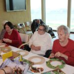 Starejše stanovalke sede ob mizi izbirajo okraske in jih nameščajo na praznične venčke, pred njimi so škatle z materialom in osnutki venčkov.