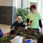 Stanovalka in animatorka skupaj ustvarjata praznični aranžma iz vejic, storžev in sveč; na mizi je razporejen ustvarjalni material.