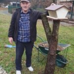 Stanovalec doma za starejše stoji ob drevesu in drži leseno ptičjo hišico, pripravljeno za namestitev.