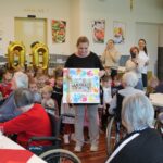 Skupina otrok in vzgojiteljic stoji pred stanovalci doma, v ozadju so velike zlate balonske številke 100; vzgojiteljica v rokah drži barvit plakat z otroškimi odtisi dlani.