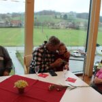 Starejši moški sedi ob mizi z majhno deklico, ki jo objame; deklica mu izroča cvetlico, ob njima sedijo še drugi stanovalci in se prijazno nasmihajo.