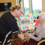 Vzgojiteljica podarja stanovalki plakat z otroškimi odtisi dlani in fotografijo skupine otrok; stanovalka se nasmejano zahvaljuje.