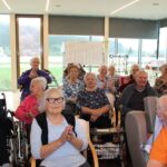 Skupina stanovalcev doma sedi v dvorani, ploska in se smehlja med praznovanjem; v ozadju je tabla za tombolo.