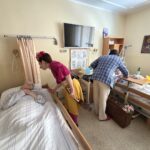 Dva bolnišnična klovna obiskujeta dve stanovalki, ki ležita v posteljah v domu starejših; eden od klovnov se sklanja k prvi stanovalki, drugi pa se pogovarja z drugo.