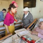 Bolnišnična klovnesa se pogovarja s stanovalcem, ki stoji ob postelji stanovalke; v ozadju drugi klovn obiskuje osebo v postelji.