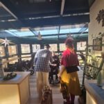 Bolnišnična klovna obiskujeta skupino stanovalcev, ki sedijo v invalidskih vozičkih v zimski terasi doma; klovnesa stoji pred njimi.