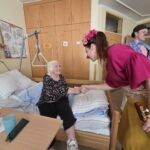 Klovnesa s rdečim nosom se rokuje s stanovalko, ki sedi na robu postelje; drugi klovn stoji v ozadju in opazuje.