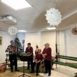 Skupina glasbenikov v ubranih bordo oblačilih stoji pred občinstvom, medtem ko moški v ospredju igra na harmoniko.