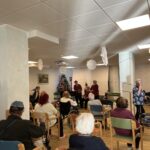 Stanovalci sedijo v polkrogu in spremljajo nastop glasbenikov na odru, v ozadju pa je okrašena smrečica.