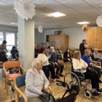 Občinstvo starejših oseb sedi na stolih in invalidskih vozičkih ter opazuje glasbeni nastop na drugi strani prostora.