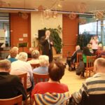 Moški z mikrofonom nastopa pred številnimi starejšimi poslušalci, ki sedijo v dvorani doma starejših.