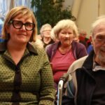 Ženska in starejši moški sedita v prvi vrsti med občinstvom in gledata proti odru.