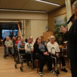 Govorec z mikrofonom nagovarja starejše udeležence, ki sedijo v vrstah in pozorno poslušajo.