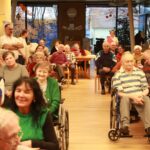 Številni stanovalci doma starejših sedijo v dvorani, obrnjeni proti odru, kjer poteka dogodek.