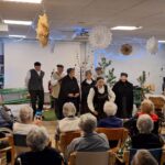 Skupina folkloristov v tradicionalnih oblačilih nastopa v praznično okrašenem prostoru doma, pred njimi sedi občinstvo stanovalcev na stolih in invalidskih vozičkih.