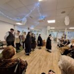 Folklorna skupina stoji v polkrogu in nastopa pred stanovalci doma; prostor je okrašen z lučkami, papirnatimi okraski in božičnimi drevesci.