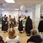 Folkloristka v tradicionalni noši med nastopom dvigne košaro, ob njej stojijo drugi člani skupine z glasbilom, stanovalci spremljajo dogajanje iz sedežev.