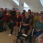 Stanovalci doma za starejše sedijo na stolih in invalidskih vozičkih ter zbrano spremljajo filmsko projekcijo v skupnem prostoru.