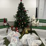Prostor z božično okrašeno jelko ter številnimi darilnimi vrečkami, razporejenimi po tleh, označenimi s številkami za srečelov.
