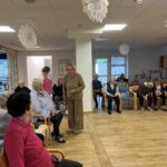 Skupina stanovalcev doma za starejše sedi v krogu, animatorka v stoječem položaju vodi družabno dejavnost.