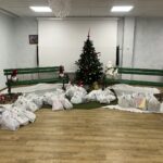 Božično okrašen prostor s smrečico in darilnimi vrečkami za srečelov, razporejenimi v polkrogu na tleh.