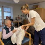 Negovalka se nasmejano sklanja k stanovalki in ji izroča dve veliki darilni vrečki med srečelovom.