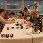 Stanovalke ustvarjajo naravna mila in sveče; na mizi so razporejeni pisani lončki s svečami in pripomočki za delo.