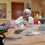 Stanovalka sedi za mizo in zbrane papirnate izrezke natančno zapisuje oziroma oblikuje pri ustvarjalni dejavnosti.