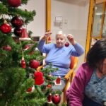 Stanovalka okrašuje božično drevesce in v roki drži rdeč božični okrasek.