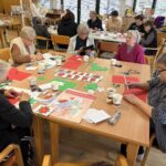 Skupina starejših oseb sedi za mizo in ustvarja praznične voščilnice iz barvnega papirja, škarij in lepila.