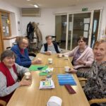 Pet starejših žensk sedi za mizo, pije kavo in se pogovarja med izdelovanjem voščilnic.