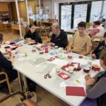 Starejši udeleženci in mladi prostovoljci skupaj ustvarjajo praznične voščilnice iz rdečega in belega papirja.